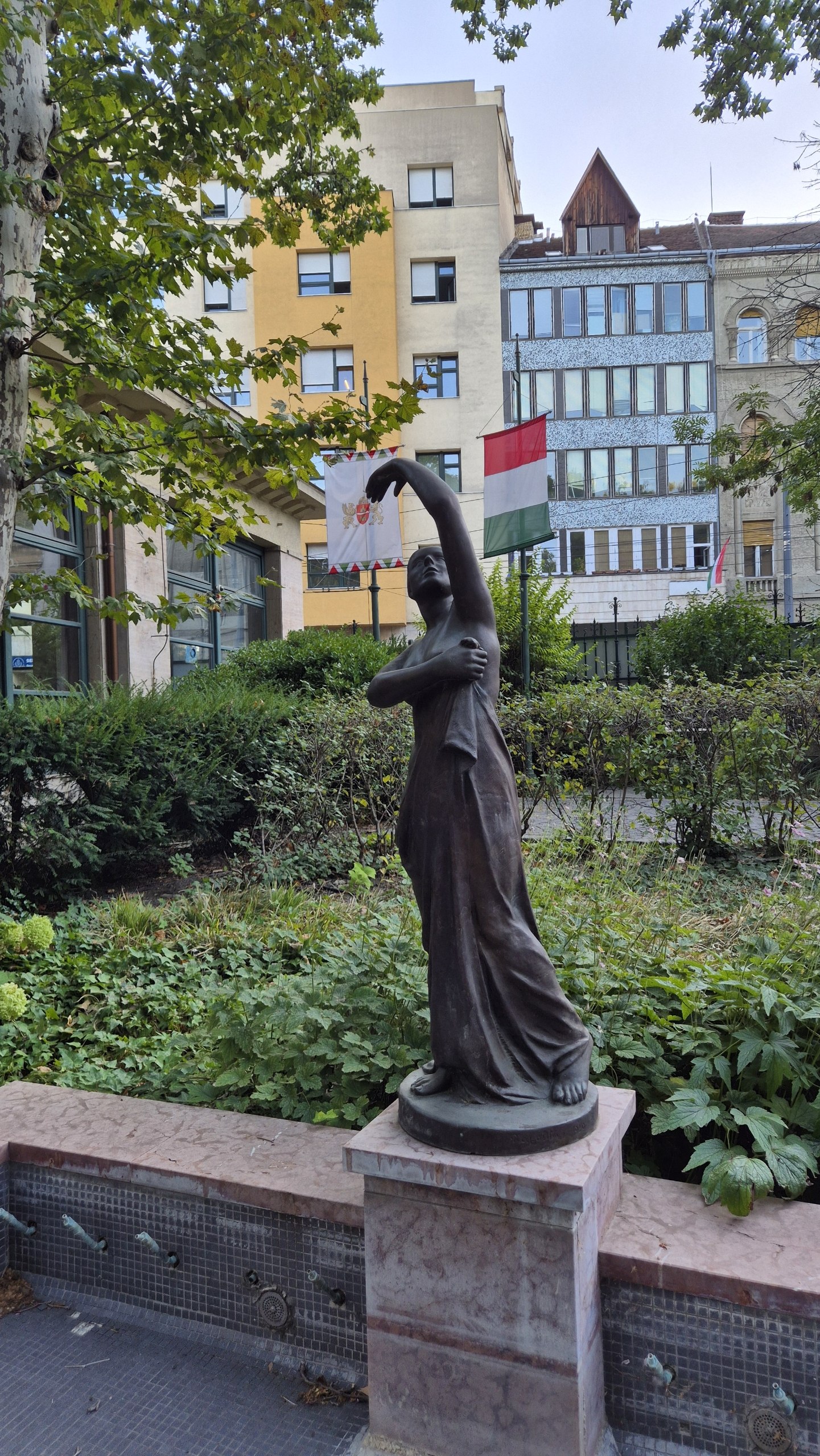 A bronze female figure in the garden at Lukács Baths with a Hungarian flag behind it