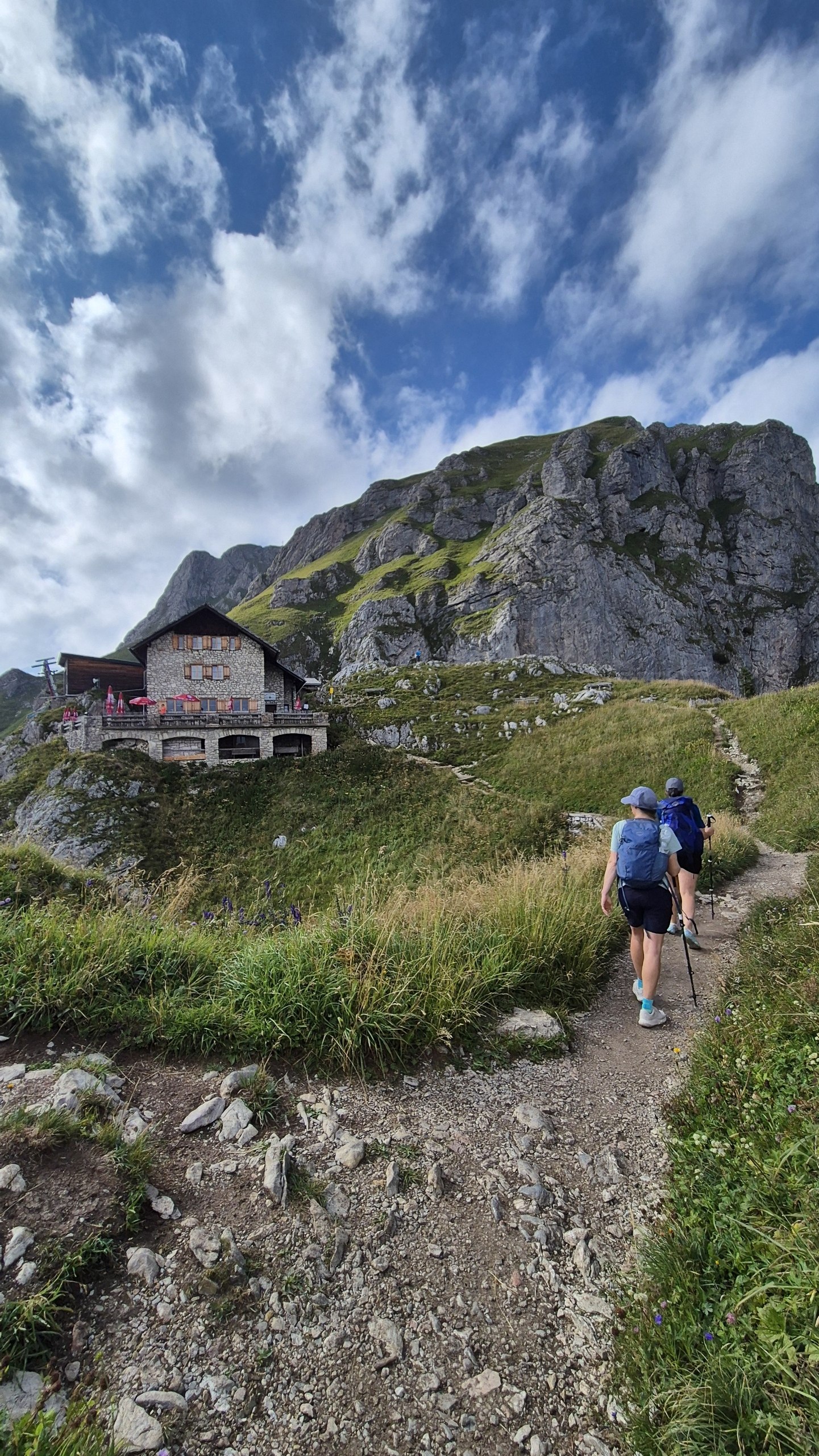 Hikers approaching a stone mountain hut beneath rocky cliffs